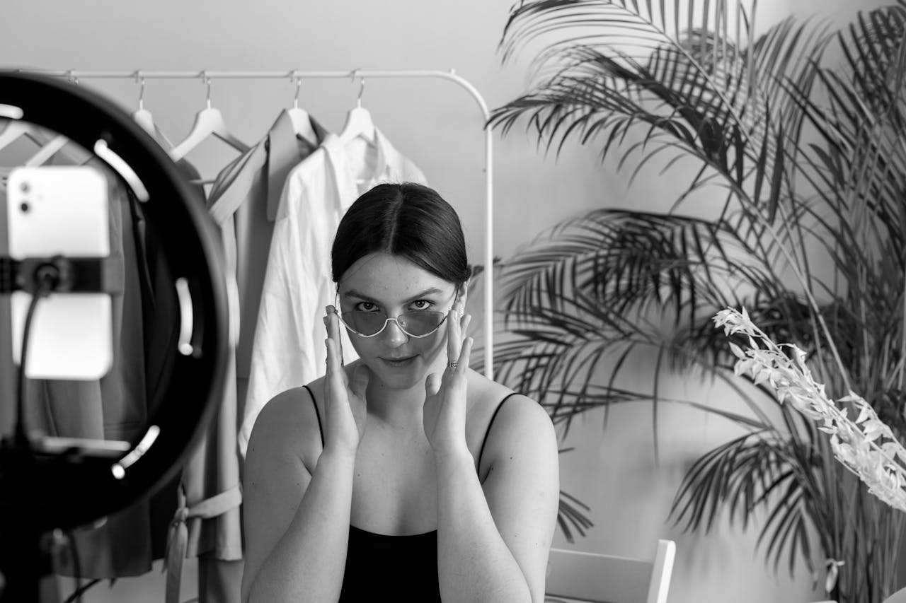 Young woman in stylish clothing filming a fashion vlog indoors with a smartphone and ring light, surrounded by plants.